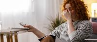 Stressed woman sitting on a couch rubbing her eyes while holding glasses and a notebook at home