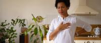 Woman standing in kitchen holding her chest and lower back, appearing to experience chest pain or discomfort at home