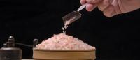 Hand pouring pink Himalayan salt crystals into a wooden bowl, with a salt grinder nearby.
