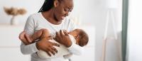 A young African-American woman holding a newborn baby in her arms. The woman is wearing a white long-sleeved shirt and blue jeans. She has long braided hair and is smiling at the baby. The baby is wrapped in a white blanket and is sleeping peacefully.