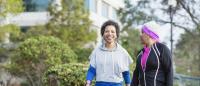 Two African-American women walking hand in hand on a sidewalk. They are both wearing workout clothes and appear to be in a park or garden. The woman on the left is wearing a gray hoodie and black leggings, while the woman in the middle has a pink headband
