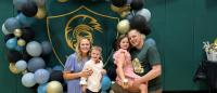 A family of four posing for a photo in front of a large balloon arch. The arch is made up of blue, black, and gold balloons, with a green and gold logo in the center. The family consists of a man, a woman, and two young children.