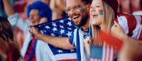 A young couple, a man and a woman, sitting in a stadium and holding American flags. They are both smiling and appear to be happy. The man is wearing glasses and has a beard, while the woman has her face painted in the colors of the American flag.