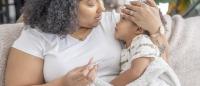 A mother and her young daughter sitting on a beige couch. The mother is wearing a white t-shirt and has curly hair. She is holding a thermometer in her hand and is looking down at the daughter with a concerned expression on her face.