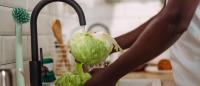 A person's hands washing a large head of cabbage in a kitchen sink. The person is wearing a white t-shirt and is standing in front of a white countertop with a black faucet.