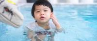 A young child in a swimming pool. The child is in the center of the image, with their body partially submerged in the water. They are wearing a white swimsuit with red and white polka dots. Their hand is on their ear.