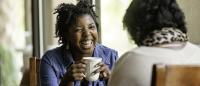 Two women sitting at a table in a cafe or restaurant. The woman on the left is African-American and is wearing a denim jacket. She has curly hair and is holding a white coffee mug in her hands and is smiling widely.