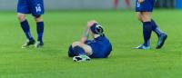 A soccer player lying on the ground with his head in his hands. He is wearing a blue jersey and white soccer cleats. His body is covered in sweat and his arms are stretched out to the sides.
