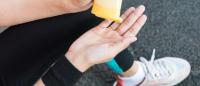 A close-up of a person's hands holding a yellow water bottle. The person is wearing black leggings and white sneakers. The background is blurred, but it appears to be an outdoor setting with a concrete floor.