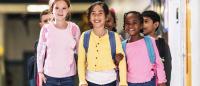 A group of six young children walking in a hallway. They are all wearing backpacks and are smiling at the camera. The girl in the center is wearing a yellow cardigan and blue jeans, while the girl on the left has blonde hair and carries a pink backpac