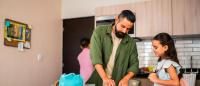 A man and a young girl in a kitchen. The man is standing in front of a round white table with a blue bag on it. He is wearing a green jacket and has a beard. The girl is standing next to him, wearing a pink backpack.