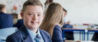 A young boy sitting at a desk in a classroom. He is wearing a blue suit and a red tie. He has short blonde hair and is smiling at the camera. Behind him, there are other students sitting at their desks and a teacher standing in front of a whiteboard