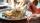 Close-up of a person eating a plate of fried rice with vegetables using a knife and fork, with natural sunlight streaming in and a blurred background of a dining setting.”